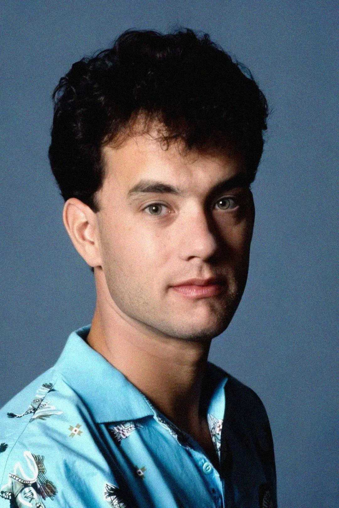 A young man with short dark curly hair and green eyes looks at the camera with a slight smile, wearing a light blue patterned shirt against a plain blue background.