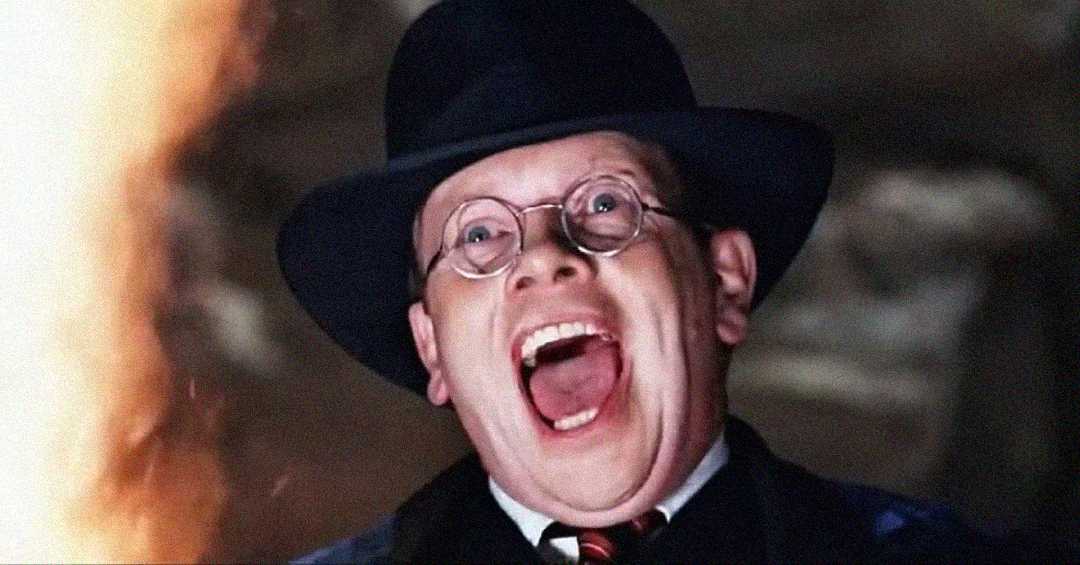 A man wearing round glasses, a dark hat, and a suit screams with his mouth wide open, looking terrified as a bright orange light or flame appears on the left side of the image.