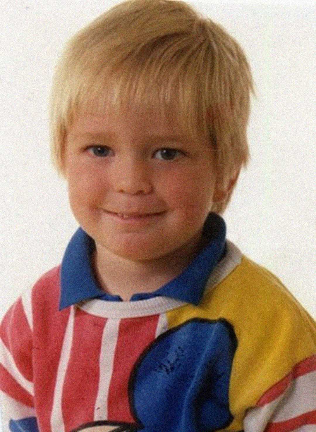A young child with blonde hair smiles slightly while wearing a colorful striped shirt with a cartoon character design on the front, posing against a plain white background.