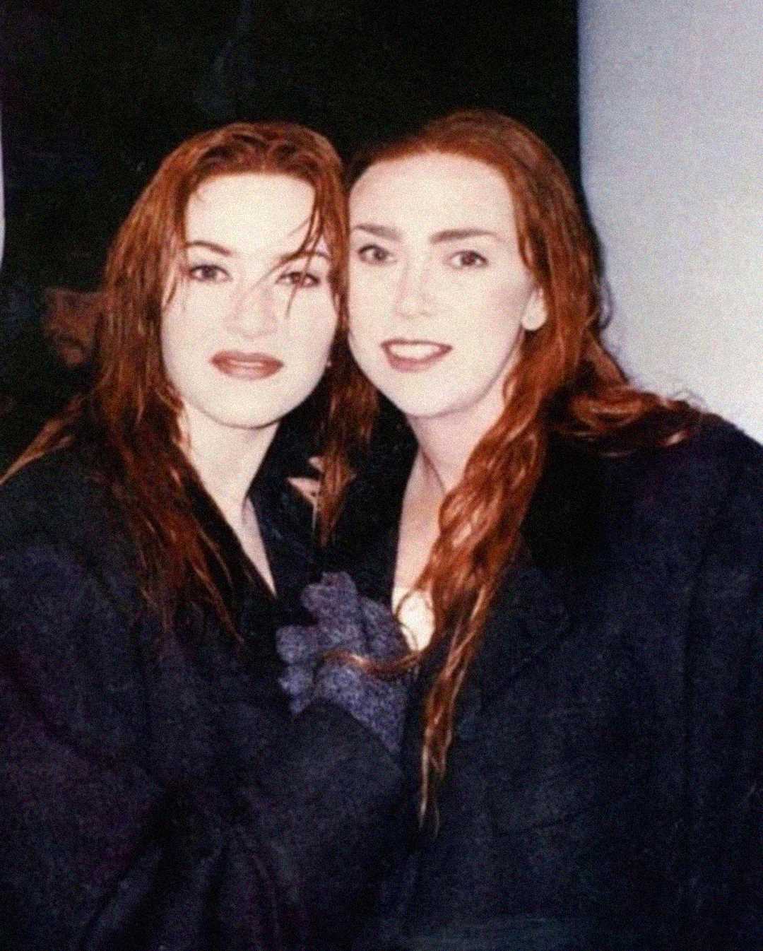 Two women with long, wet, reddish-brown hair stand close together, both wearing dark coats. They are looking at the camera and smiling slightly, appearing to be in an indoor or dimly lit setting.