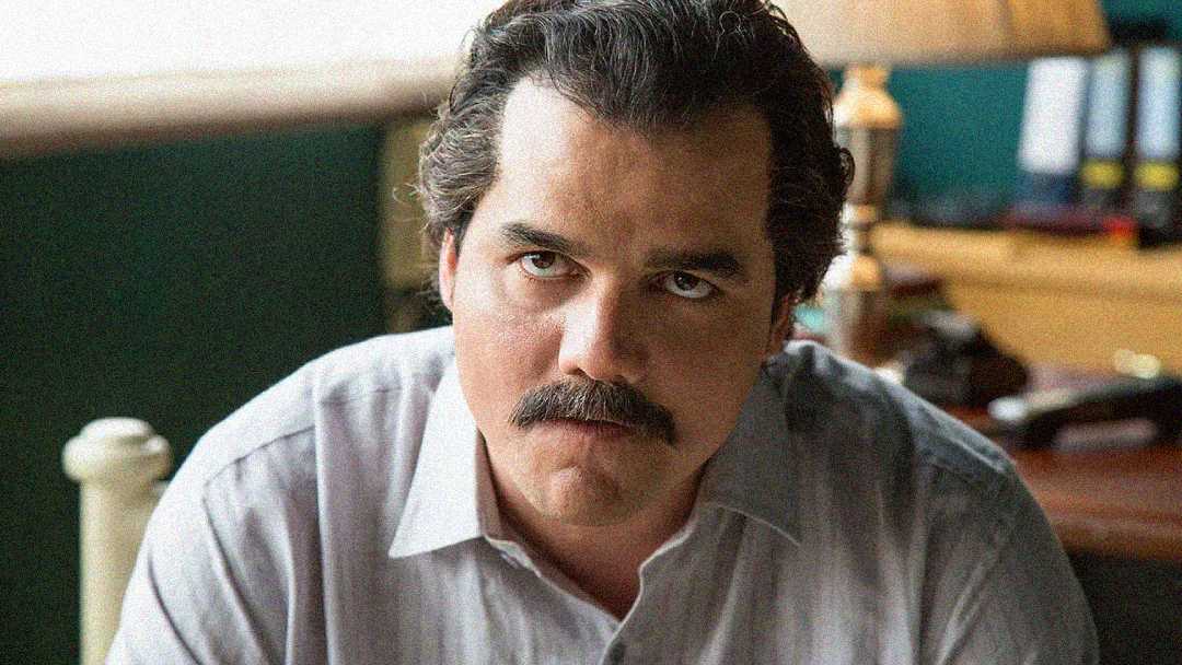 A man with dark hair and a mustache, wearing a light-colored shirt, sits indoors and looks seriously at the camera. There is a lamp and some office items blurred in the background.