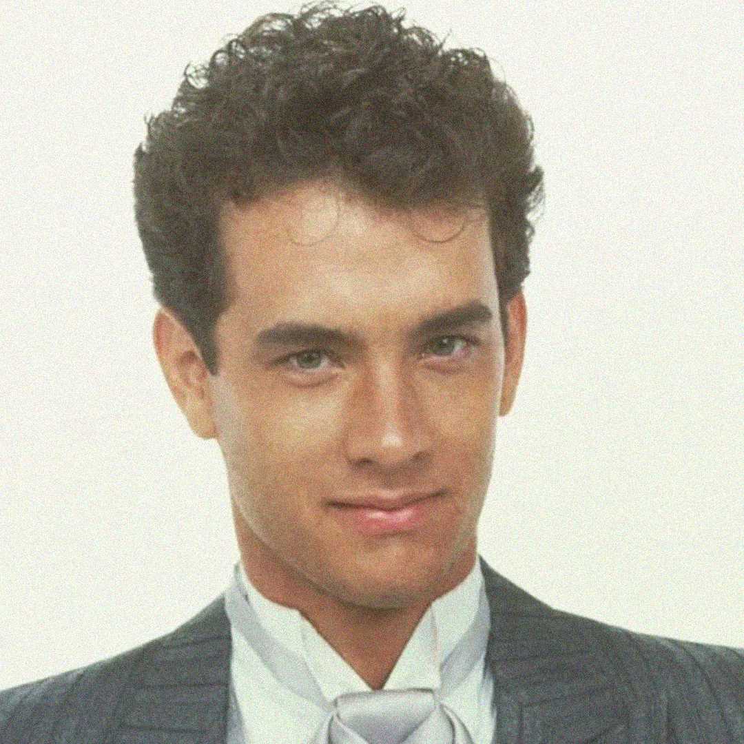 A young man with curly dark hair smiles slightly, wearing a grey pinstripe suit and a white shirt with a cravat, posing in front of a plain light background.