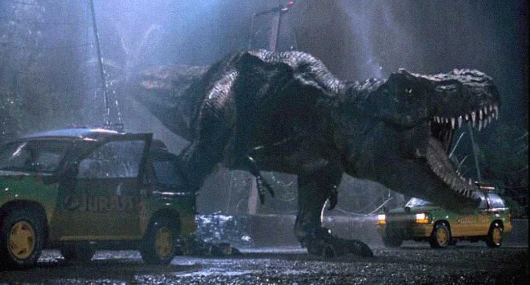 A large Tyrannosaurus rex roars at night in the rain between two damaged SUVs, one with its door open. The scene is dark and dramatic, with broken wires in the background.