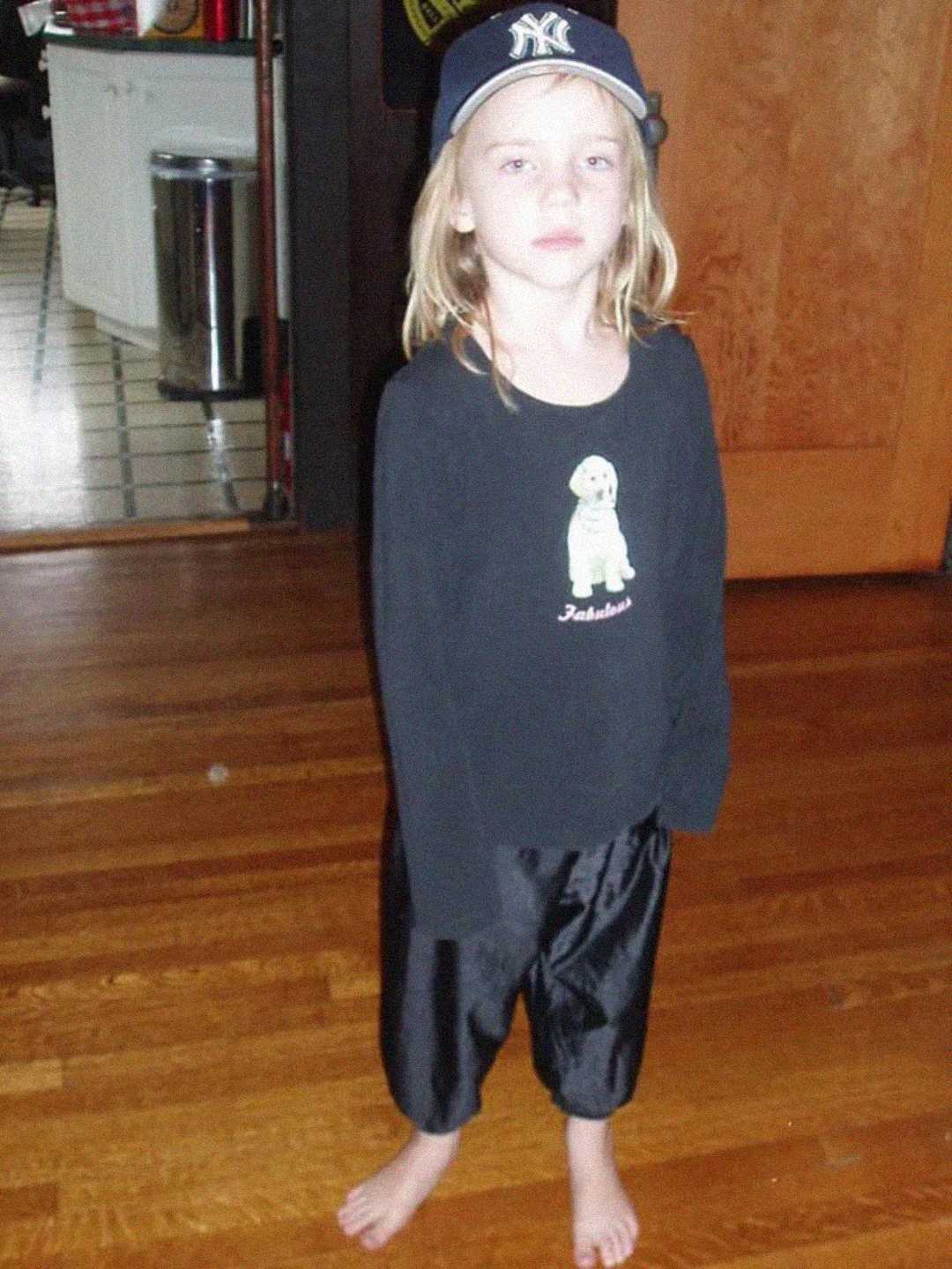 A young child stands barefoot on a wooden floor, wearing black pants, a black long-sleeve shirt with a cartoon figure, and a blue New York Yankees cap backwards. The child has long blonde hair and a neutral expression.