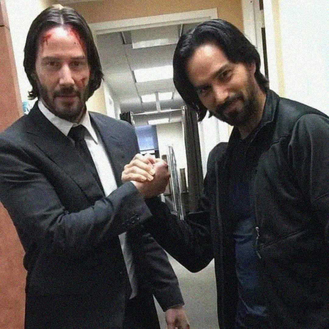 Two men clasp hands in a hallway. The man on the left wears a suit and has fake blood on his forehead, while the man on the right wears a dark jacket. Both are looking at the camera and smiling slightly.