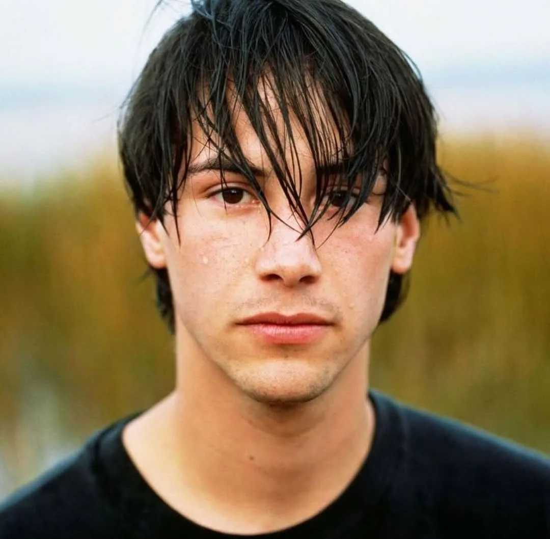 A young person with wet, dark hair falling over their face looks directly at the camera, wearing a black shirt, standing outdoors with blurred tall grass in the background.