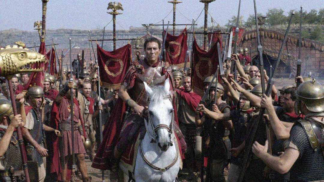 A man dressed as a Roman general rides a white horse, surrounded by soldiers in Roman armor holding red banners and spears, set in an ancient outdoor environment.