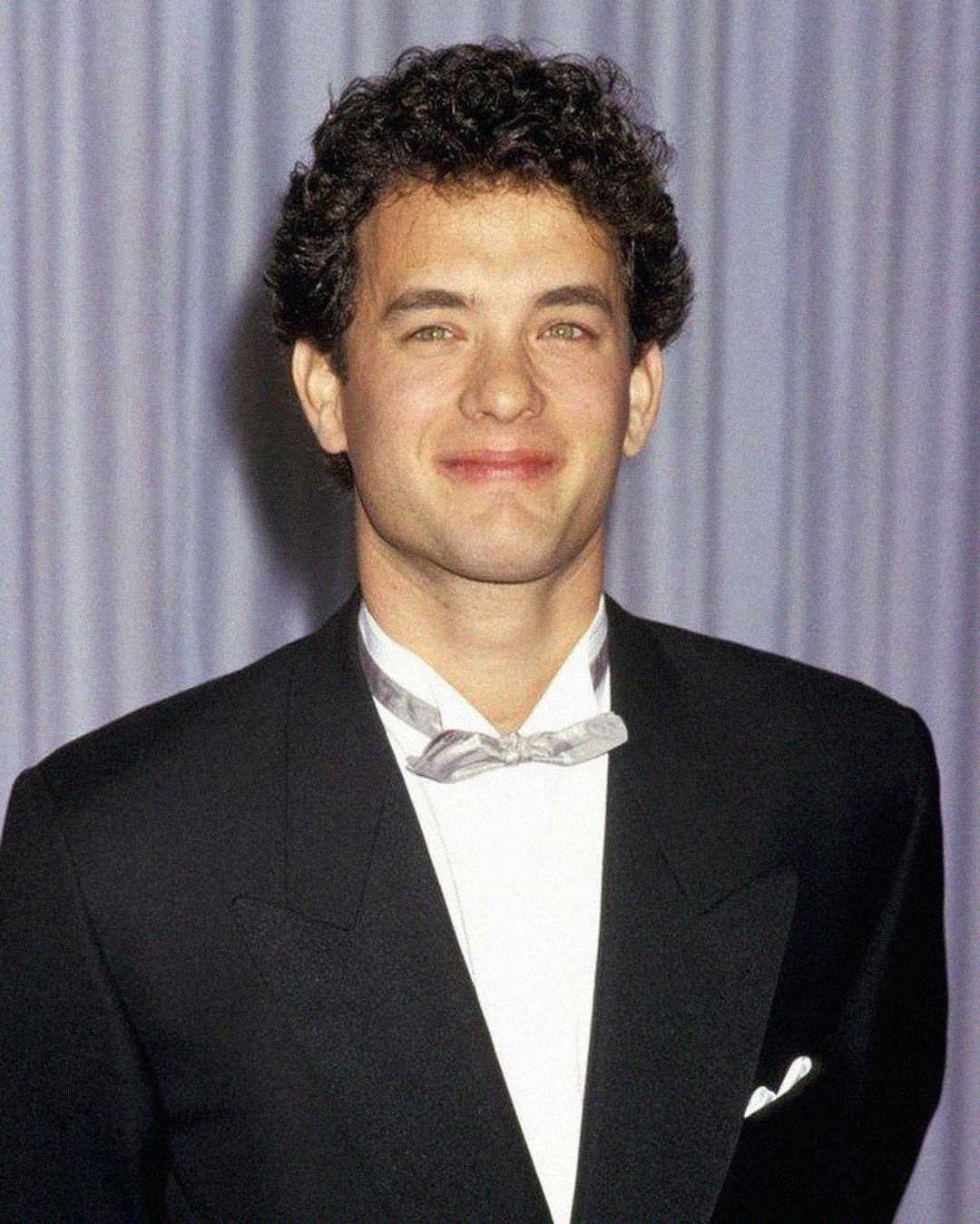 A man with short, curly dark hair is wearing a black tuxedo jacket, white shirt, and silver bow tie, standing in front of a light, pleated curtain backdrop. He is smiling slightly at the camera.