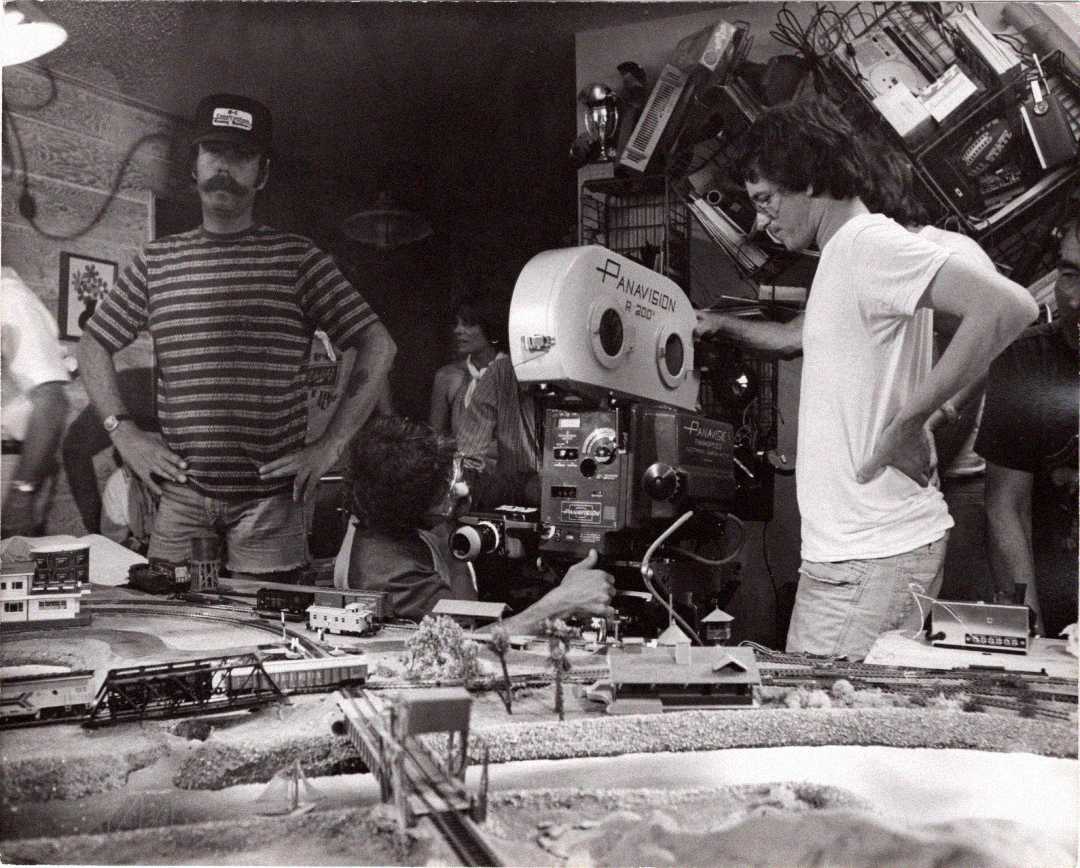 Black-and-white photo of a film crew on set. One person operates a large Panavision camera next to a model train set, while others stand nearby. Equipment and props fill the busy room.