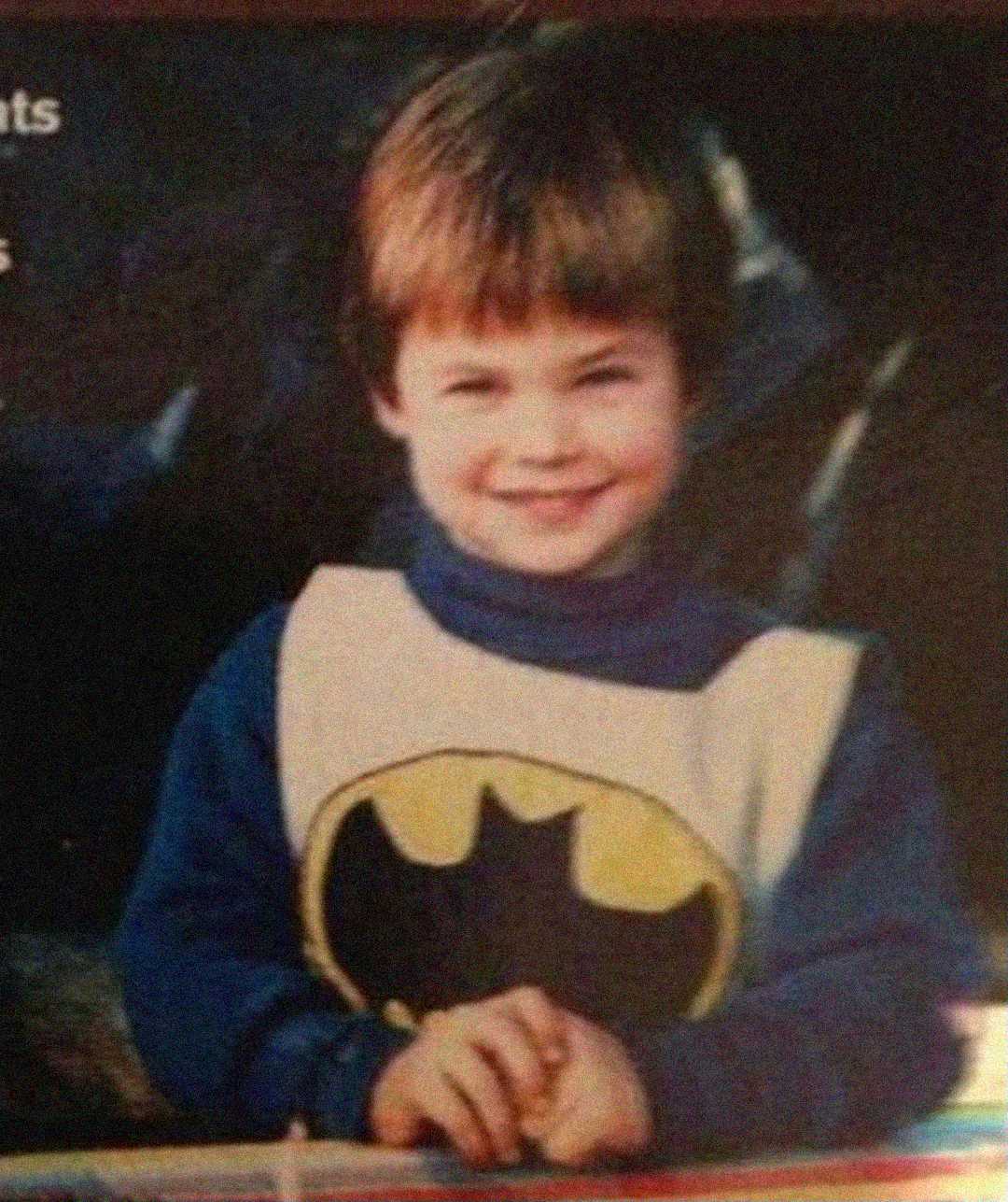 A young child with short brown hair smiles, wearing a blue shirt and a light vest with the Batman logo on the front.
