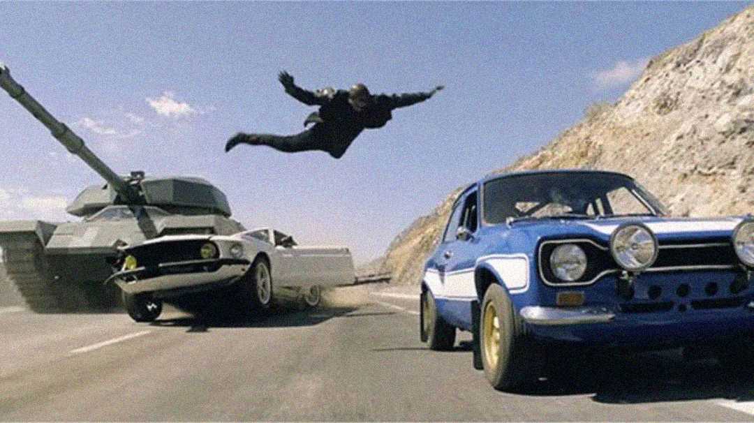 A man leaps dramatically through the air between a moving tank and two speeding cars—a white car tilting sideways and a blue car—on a sunlit mountain road.