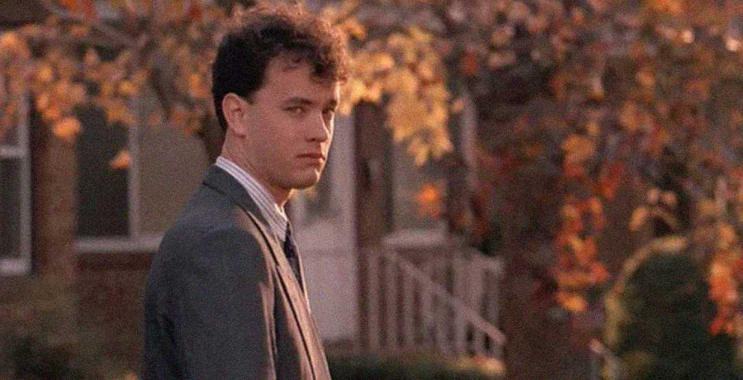 A man in a suit stands outdoors, glancing over his shoulder. The background shows autumn trees with colorful leaves and a house with a porch, giving a warm, nostalgic atmosphere.