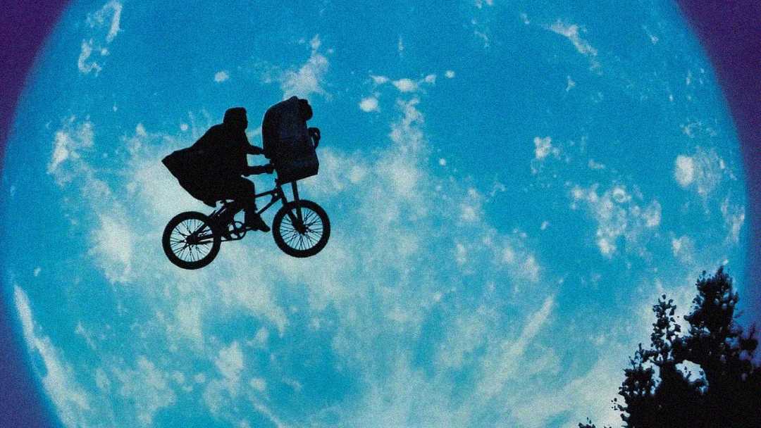 Silhouette of a person riding a bicycle with a basket, flying across a large, bright full moon in the night sky, with some trees visible at the bottom right.