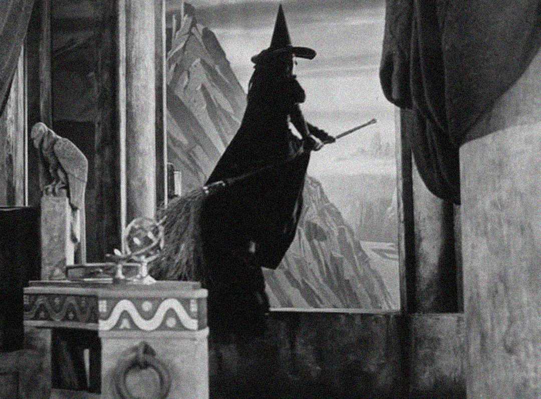 A person in a tall, pointed hat and dark cloak stands indoors near a window, holding a broomstick and looking out toward a rocky mountain landscape. The scene appears to be from a classic black-and-white film.