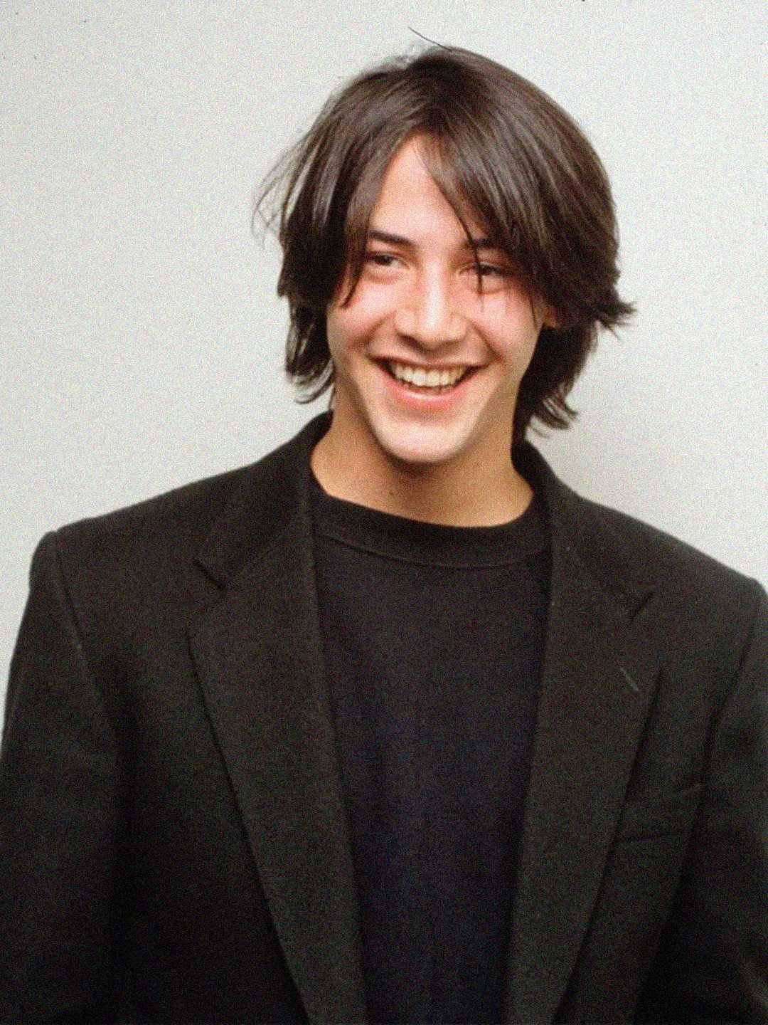 A young person with medium-length dark hair, wearing a black blazer over a black shirt, smiles brightly against a plain light background.