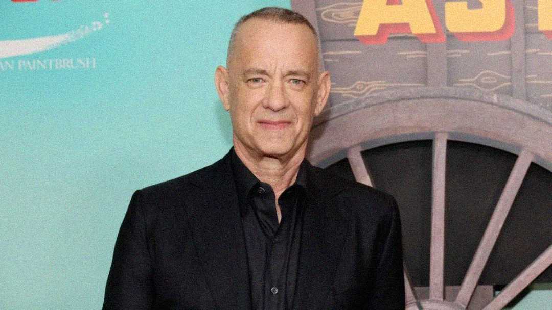 A man with short gray hair, wearing a black suit and black shirt, stands in front of a turquoise and wood-paneled background with yellow and red text partially visible.