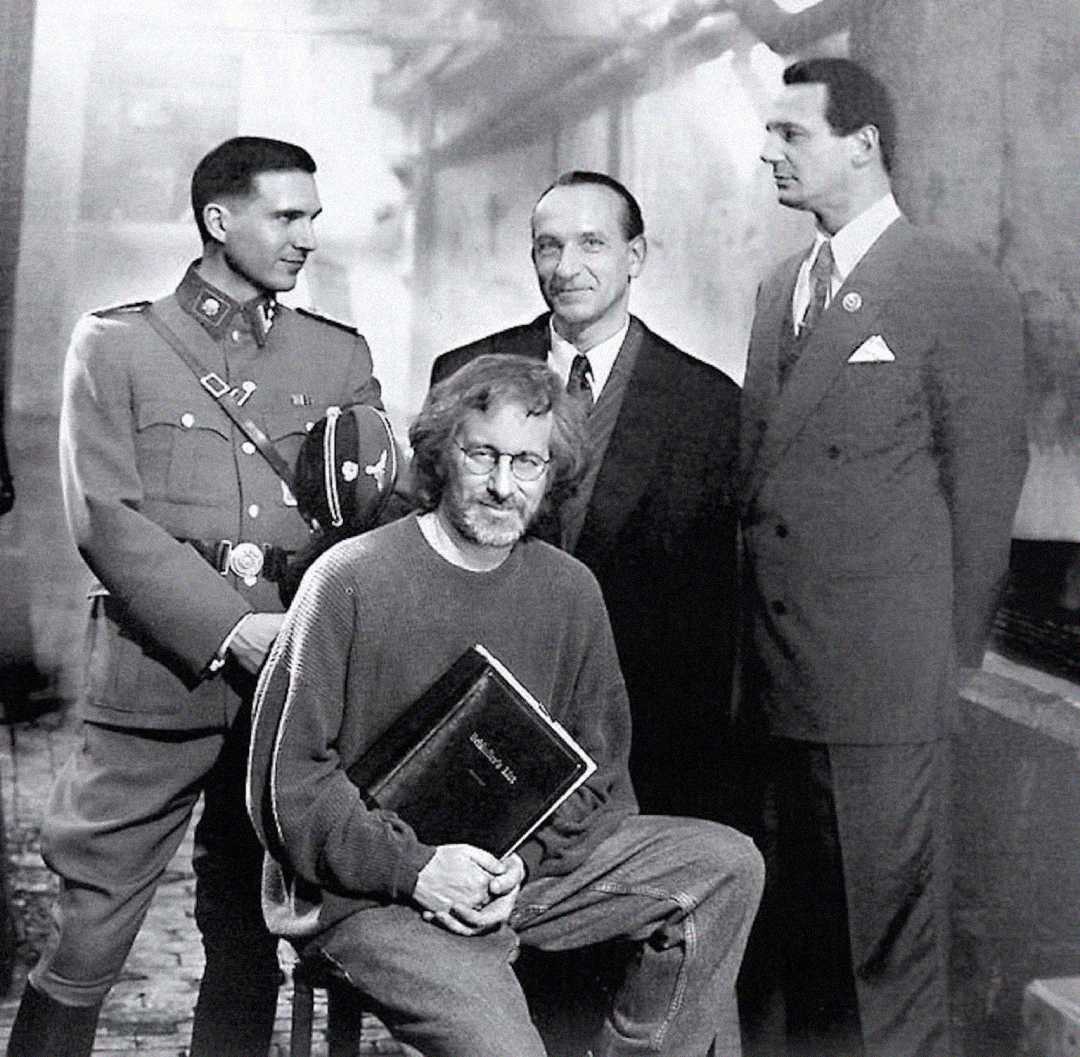 A black-and-white photo shows a man with glasses and a beard sitting on a stool holding a book, while three men in formal and military attire stand behind him, smiling. The setting appears to be a film set or historical scene.