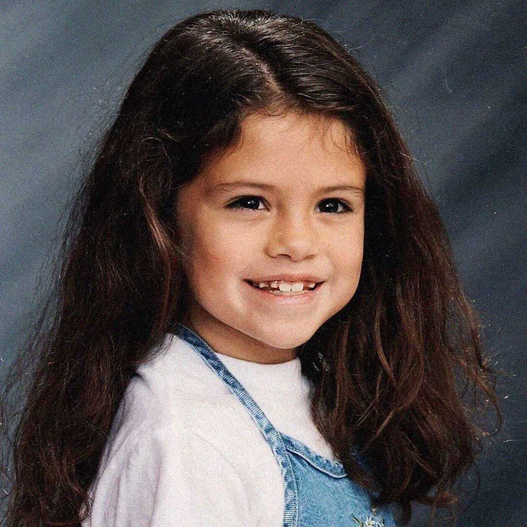 A young girl with long brown hair smiles, showing a gap where a tooth is missing. She wears a white shirt under blue denim overalls and poses in front of a softly blurred studio background.