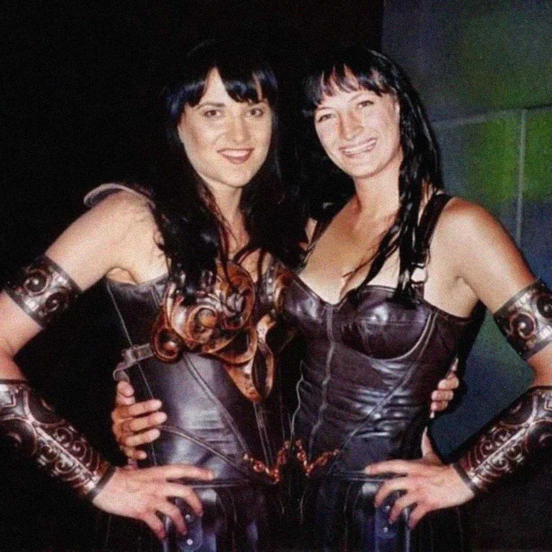 Two women dressed in matching warrior costumes with dark wigs, leather armor, and arm bracers, standing side by side with their arms around each other, smiling at the camera.