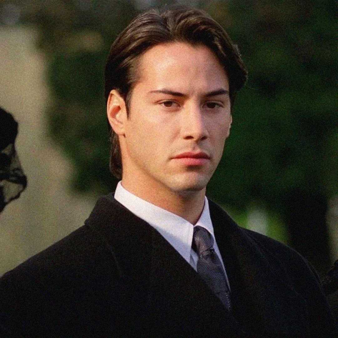 A young man with straight dark hair wearing a black suit, white shirt, and tie stands outdoors, looking serious. The background is blurred with greenery.