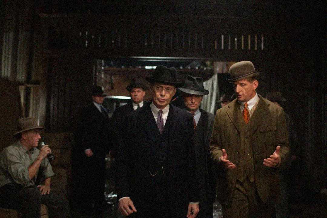 A group of men in early 20th-century suits and hats stand together in a dimly lit room; one man walks in front while another gestures with his hands, and others look on or sit in the background.