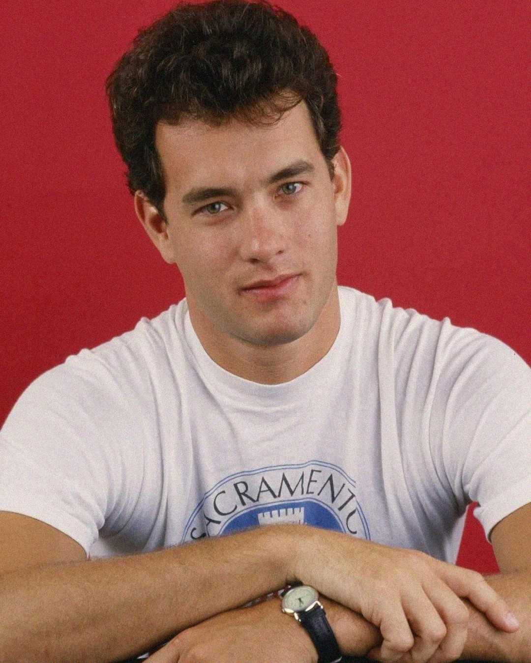 A young man with short curly dark hair, wearing a white T-shirt with a circular blue logo and a black wristwatch, sits with his arms crossed against a solid red background.