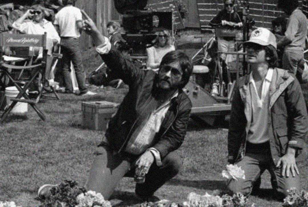 Two men on a film set, one kneeling and pointing while the other stands beside him. Film equipment, crew members, and director’s chairs are visible in the background. The image is in black and white.