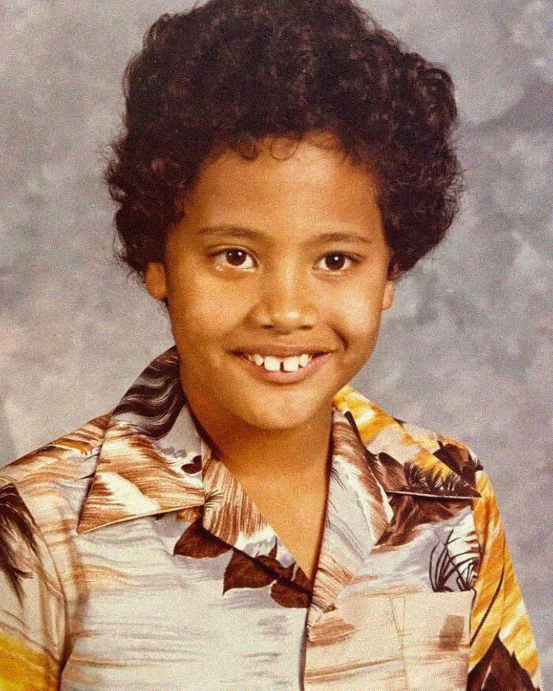 A young child with curly hair smiles at the camera, wearing a patterned button-up shirt with brown, yellow, and white colors. The background is a neutral, mottled gray.