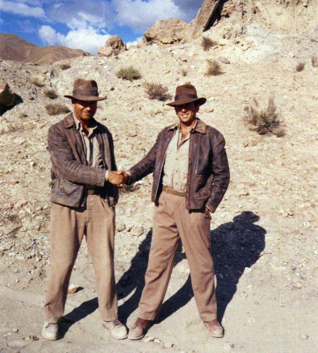 Two men wearing brown hats, leather jackets, and tan pants stand on rocky desert terrain. They are shaking hands, both smiling, with bright sunlight casting strong shadows behind them.