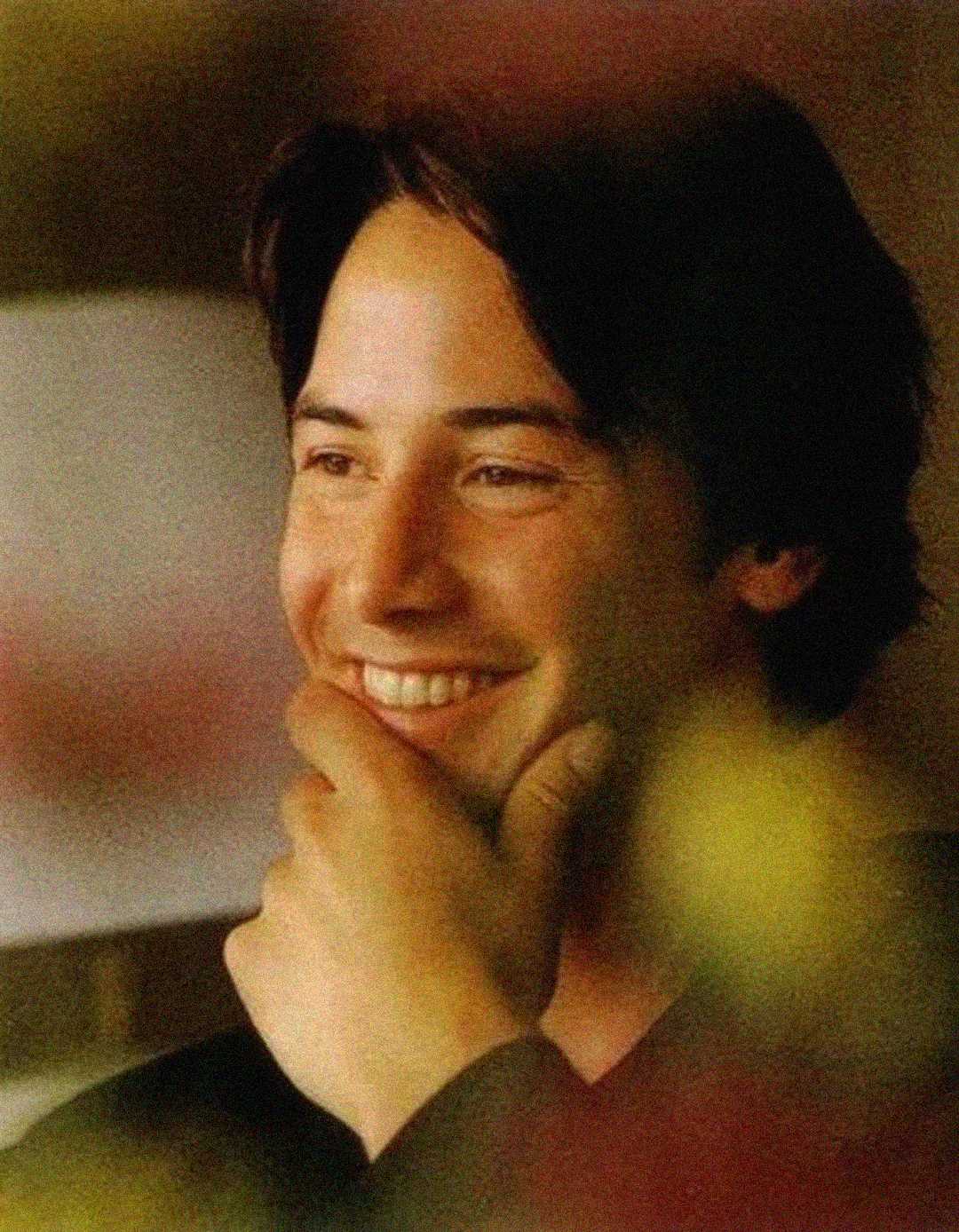 A young man with dark hair smiles warmly, resting his chin on his hand. The image is softly focused with warm, gentle lighting and blurred foreground colors, creating a cozy and relaxed atmosphere.