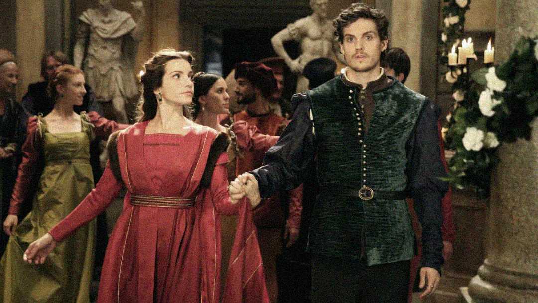 A woman in a red medieval-style dress and a man in a dark green doublet hold hands and walk together at a formal gathering, surrounded by others in period costumes.