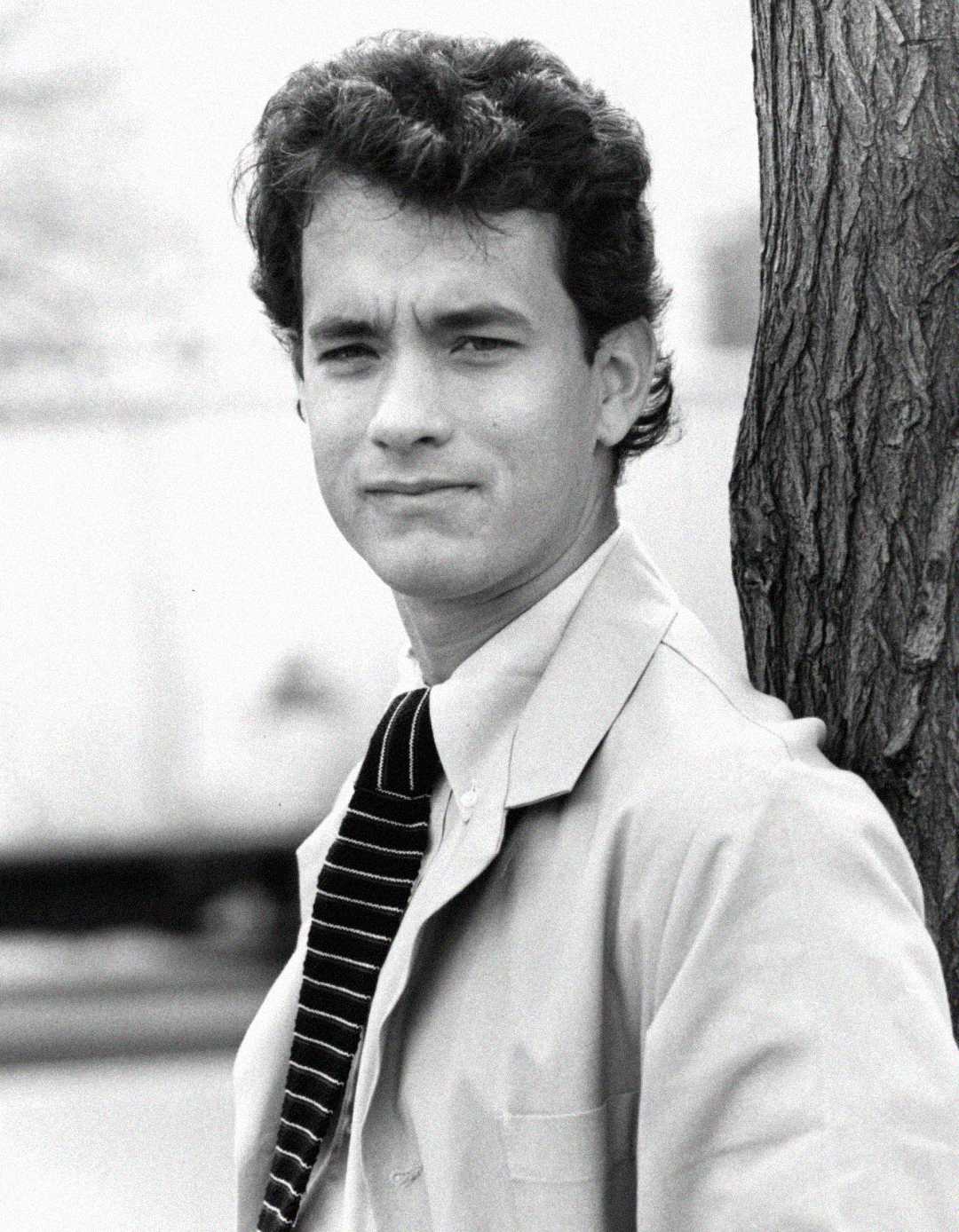 A young man in a light-colored jacket and striped tie stands outdoors next to a tree, looking at the camera with a slight smile. The image is in black and white.