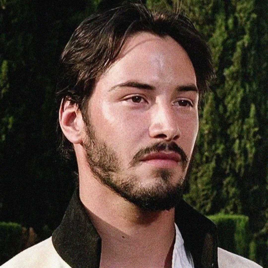 A young man with dark hair, a short beard, and a mustache stands outdoors, wearing a white shirt with a black collar. Lush green trees fill the background.