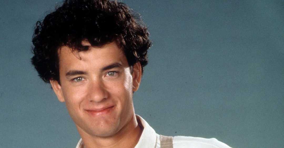 A young man with curly dark hair smiles gently at the camera, wearing a light-colored shirt and posing against a plain gray background.
