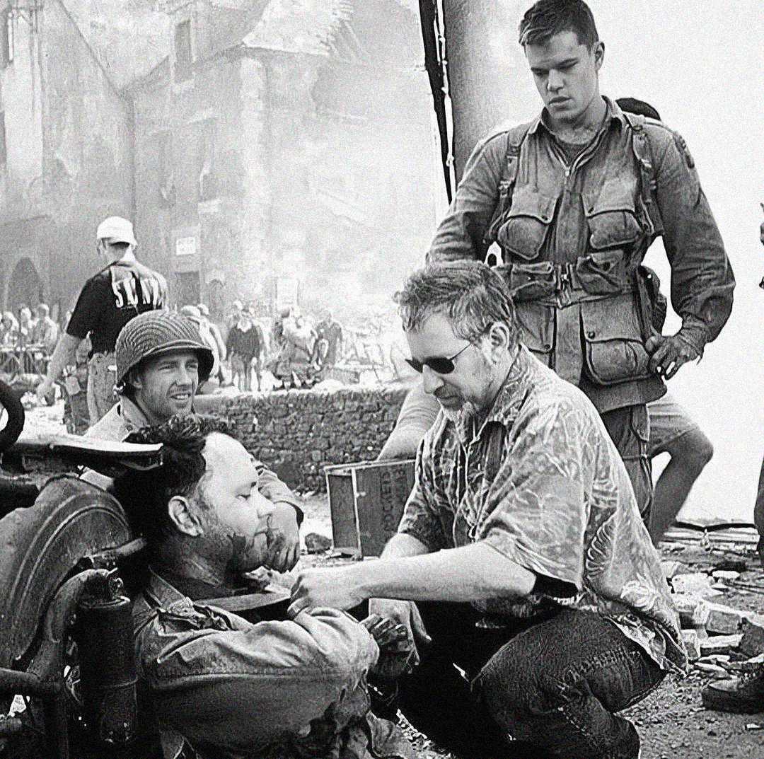 On a film set, a director kneels beside an actor in costume with fake wounds, giving directions. Other crew and actors in military gear look on, with a smoky, ruined backdrop suggesting a wartime scene.