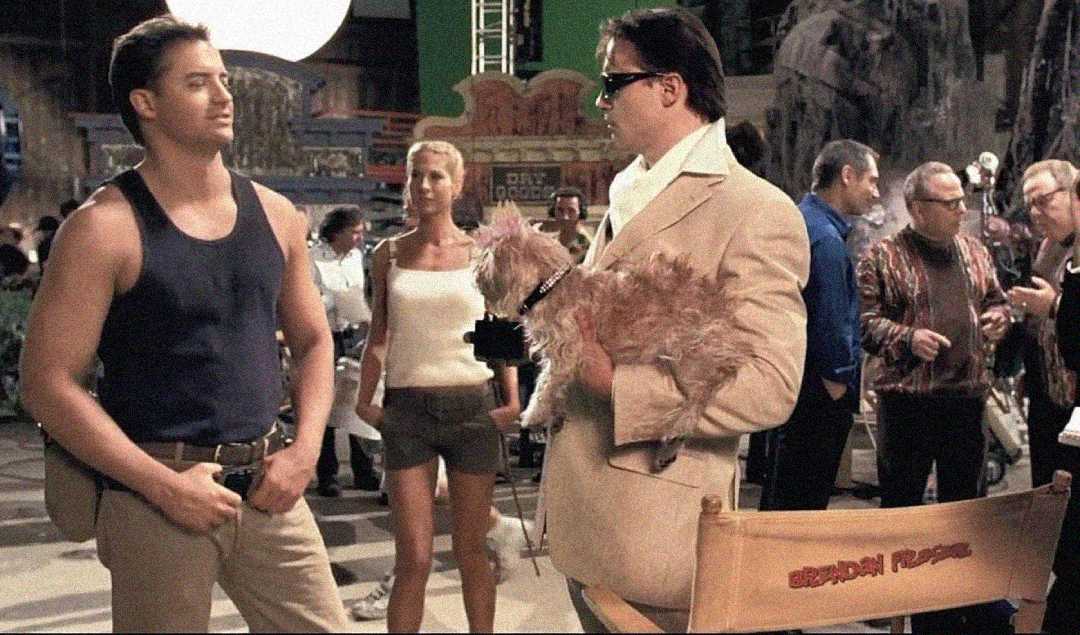 Two men face each other on a busy film set; one wears a dark tank top and jeans, the other wears sunglasses and a tan blazer while holding a small dog. Crew members and equipment are visible in the background.