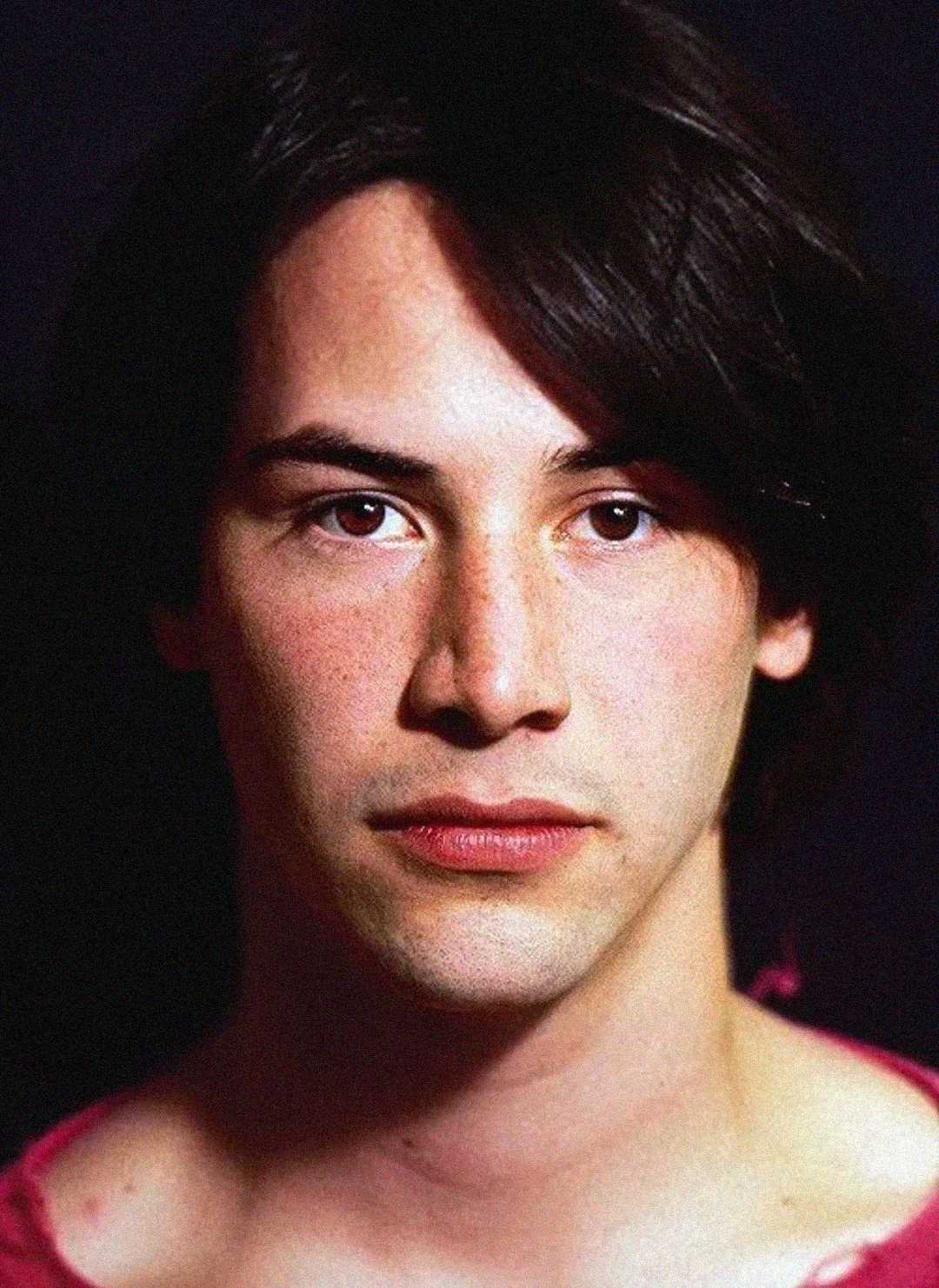 A young person with straight dark hair and fair skin, wearing a red shirt, looks directly at the camera with a neutral expression against a dark background.