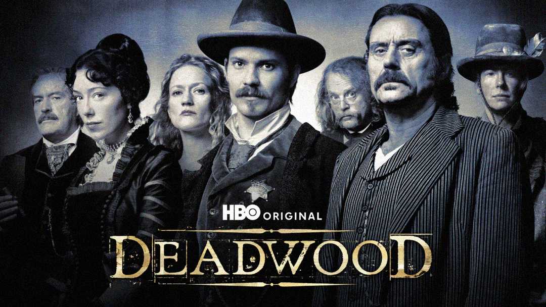A group of seven people in period Western clothing stand together, facing forward. The text “HBO Original DEADWOOD” appears at the bottom of the image. The photo has a dark, vintage tone.