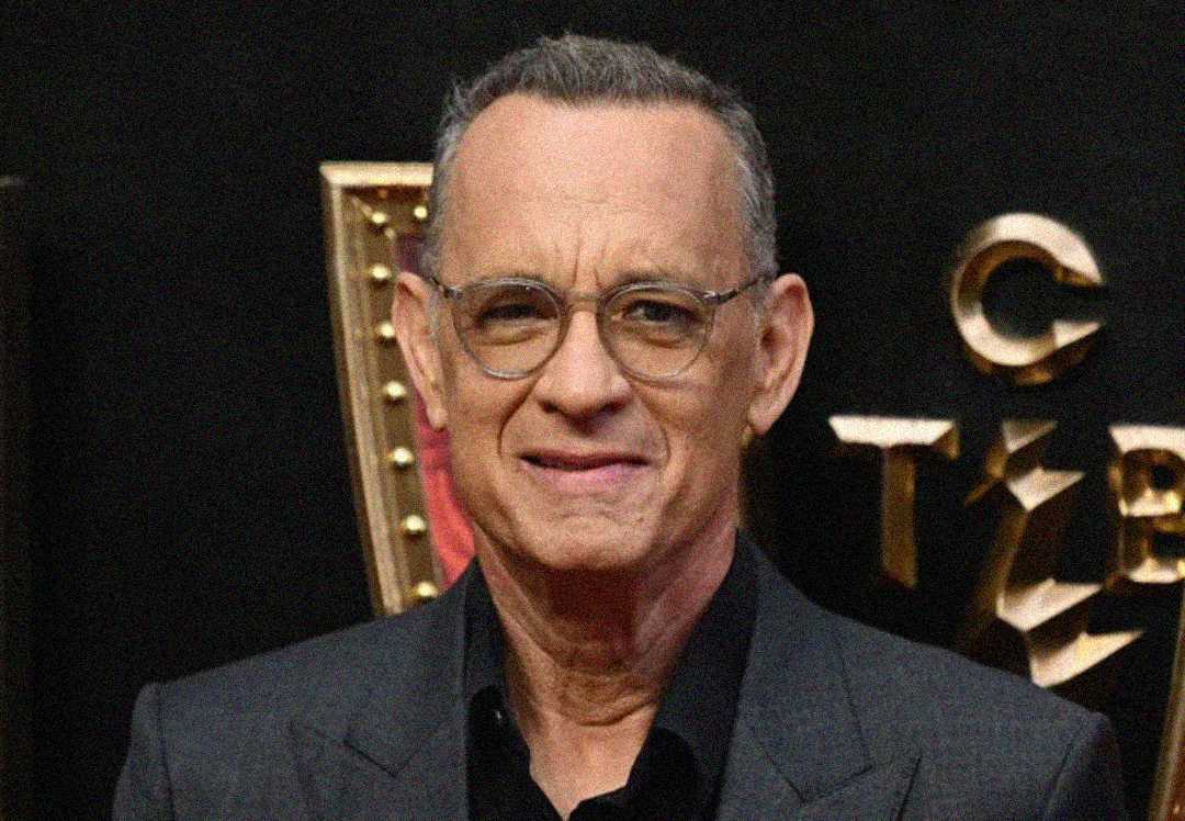 A man with short gray hair and glasses, wearing a dark suit, smiles in front of a black and gold backdrop with partially visible letters.