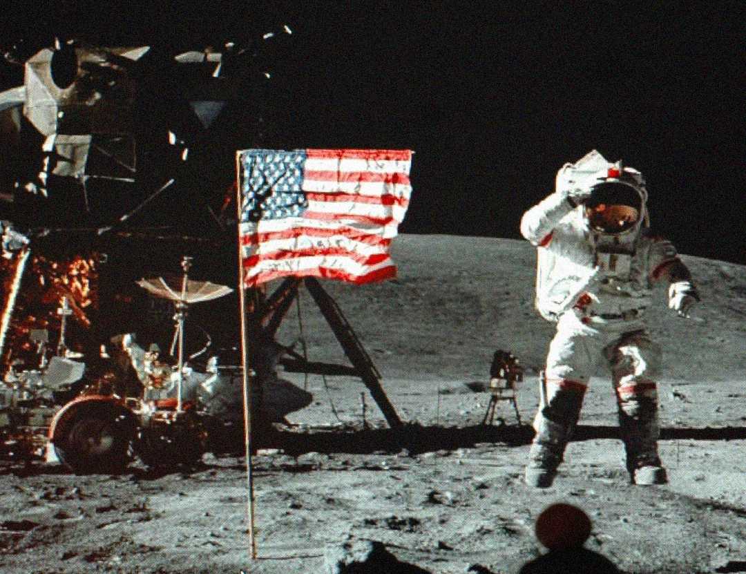 An astronaut in a white spacesuit stands on the moon beside an American flag, with the lunar module and moon rover visible in the background against a dark sky.