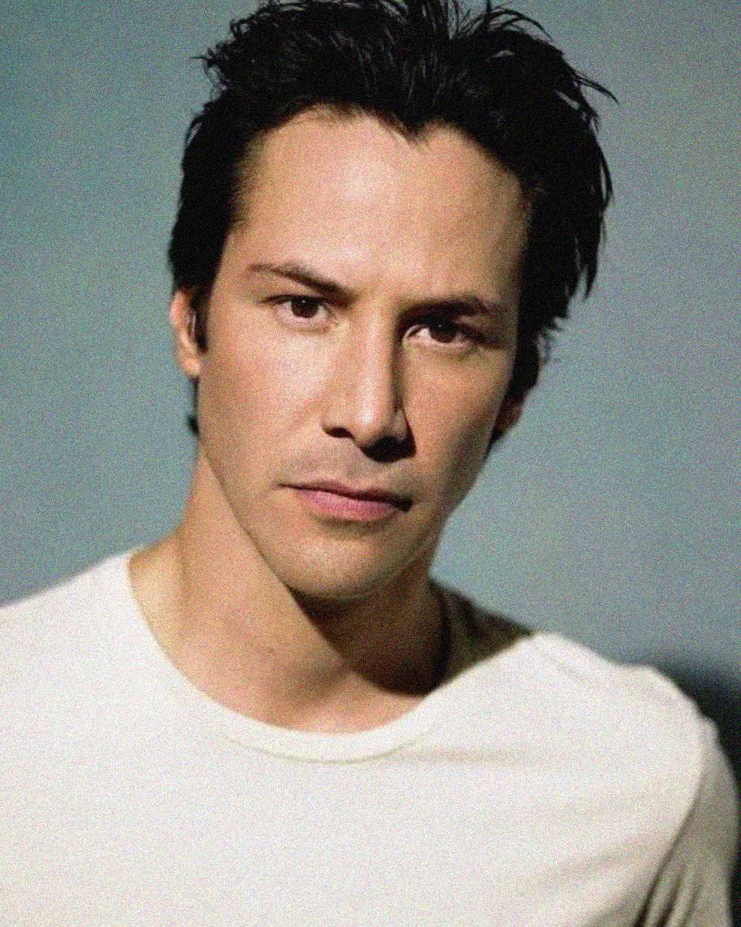 A man with short, dark hair and a serious expression is wearing a white T-shirt and looking directly at the camera against a neutral background.