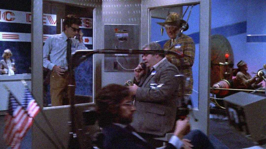 A busy newsroom scene with four men; one wears a striped suit and cowboy hat, another is seated speaking on a phone, while two others work nearby. American flags are visible in the foreground, and machines or consoles are in the background.