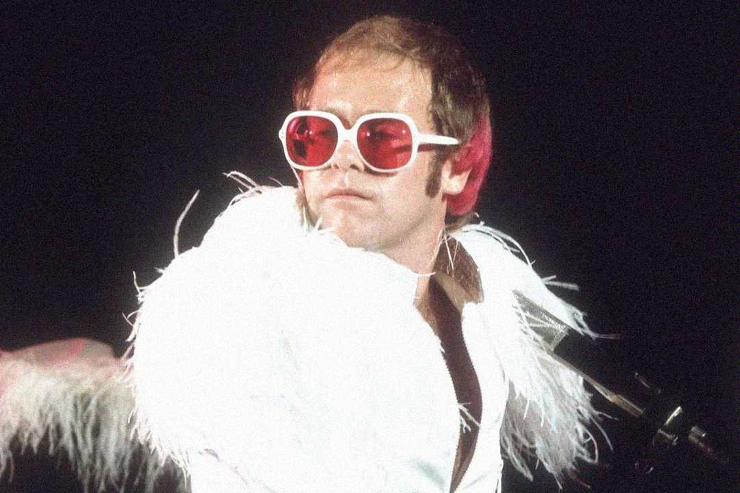 A person wearing large pink sunglasses and a flamboyant white feathered outfit performs on stage, looking to the side. The background is dark, emphasizing the dramatic costume.