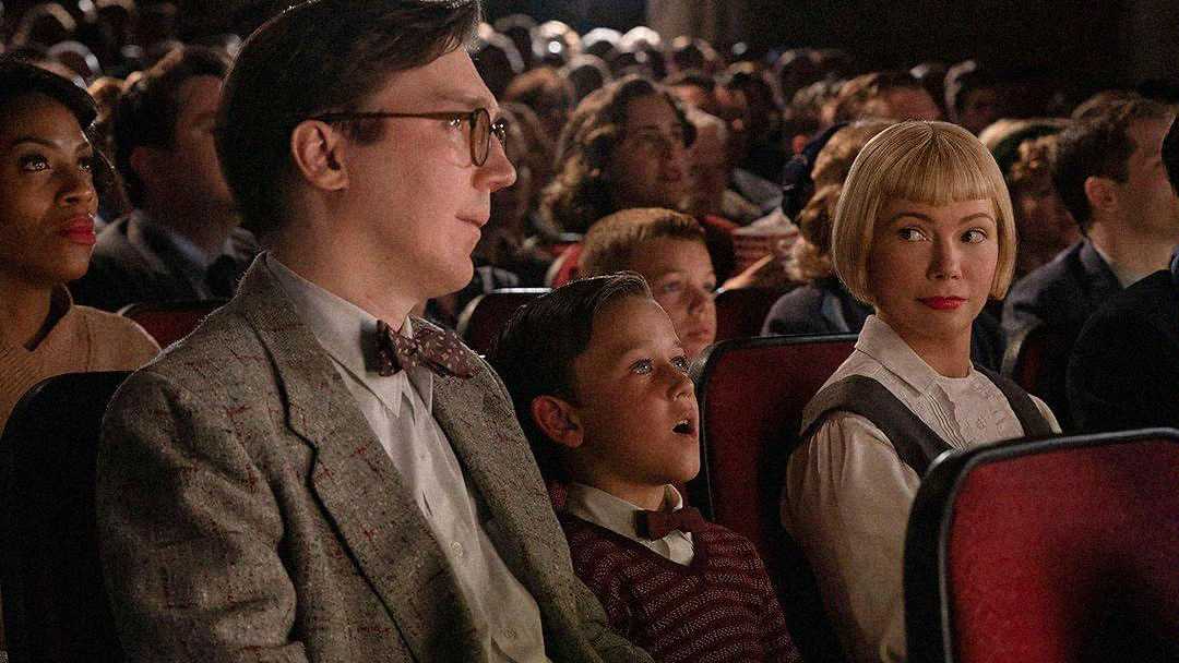 A man in glasses, a boy, and a woman with short blonde hair sit in a crowded theater, watching something on stage. The boy looks amazed, and the woman glances sideways at the man. Red seats fill the auditorium.