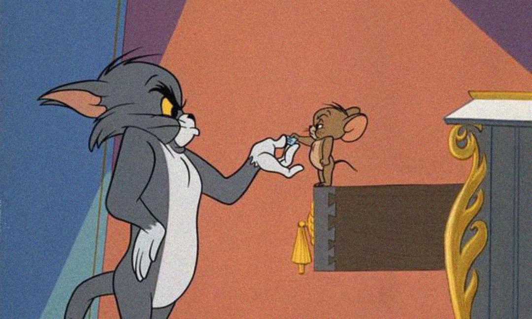 Tom, a grey cat, holds Jerry, a brown mouse, by the tail with a stern expression. Jerry looks back at Tom, standing on his tiptoes near the edge of a platform with ornate gold details.