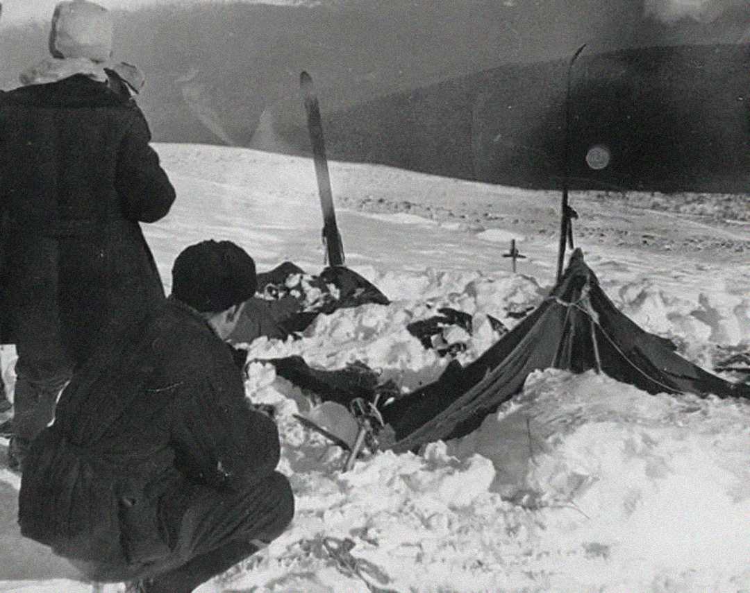 Two people in winter clothing examine a collapsed tent partially covered with snow on a snowy hillside, with a pair of skis upright and a small cross marking a spot nearby.