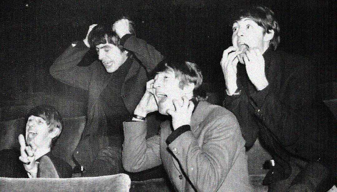 Four young men sit in theater seats, making exaggerated, playful expressions of surprise and excitement, with hands on their heads and faces. The image is black and white, suggesting it is from an earlier era.