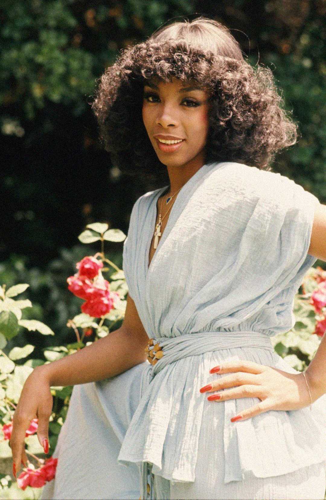 A woman with curly dark hair wearing a light blue belted dress poses in a garden with red roses, smiling at the camera with one hand on her hip.