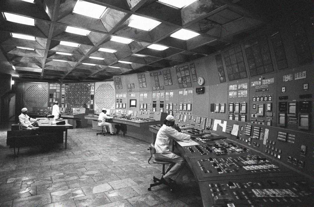 Black and white photo of people in lab coats and caps operating a large control room filled with panels, buttons, and dials, likely in a mid-20th century industrial or scientific facility.