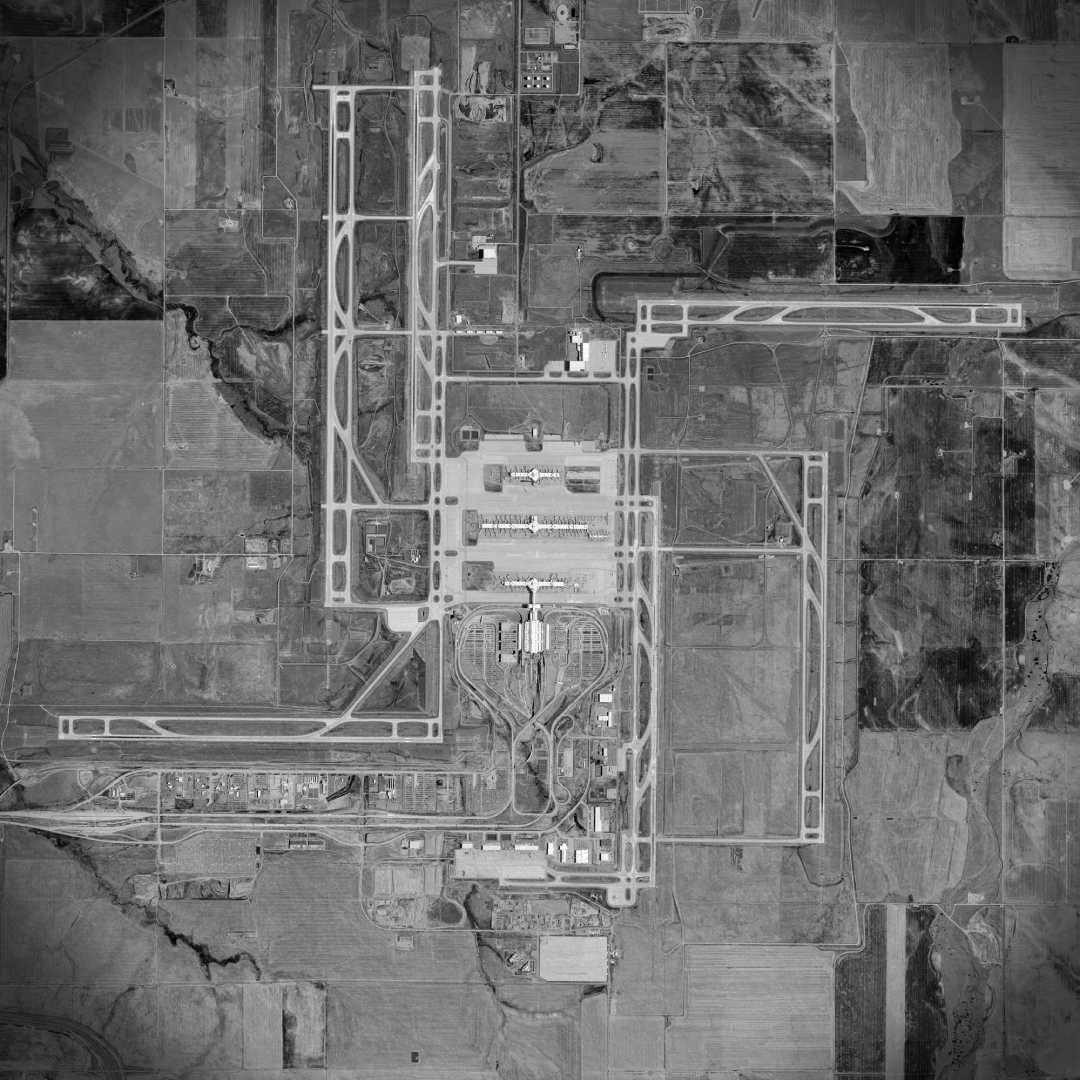 A black and white aerial view of an airport, showing runways, taxiways, terminals, and surrounding fields and roads in a grid-like pattern.